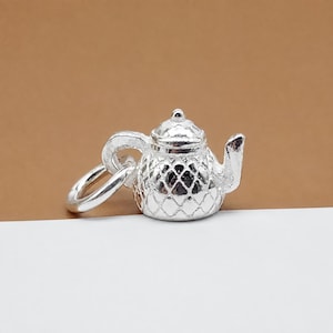 May include: A small, shiny silver-toned metal charm shaped like a miniature teapot. The teapot features a textured, diamond-patterned body, a curved handle, a spout, and a lid. A circular jump ring is attached to the handle for hanging purposes.