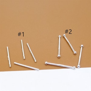 Peut inclure: Deux ensembles de clous de nez en argent. Le premier ensemble, étiqueté #1, comprend six clous avec une extrémité droite et plate. Le deuxième ensemble, étiqueté #2, comprend quatre clous avec une boule ronde à l'extrémité.