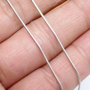 May include: Two silver snake chains, one on top of the other, are shown on a hand. The chains are thin and delicate.