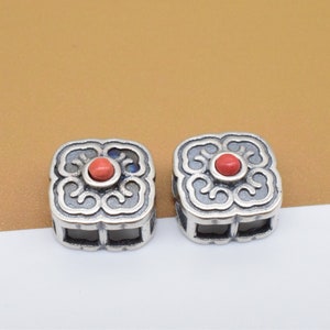 Peut inclure: Deux perles carrées argentées avec des motifs floraux complexes et une pierre précieuse rouge au centre.