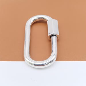 May include: A polished silver oval carabiner clasp. The clasp has a rectangular bar on one end and a hinged, curved body. The background is a light brown color.