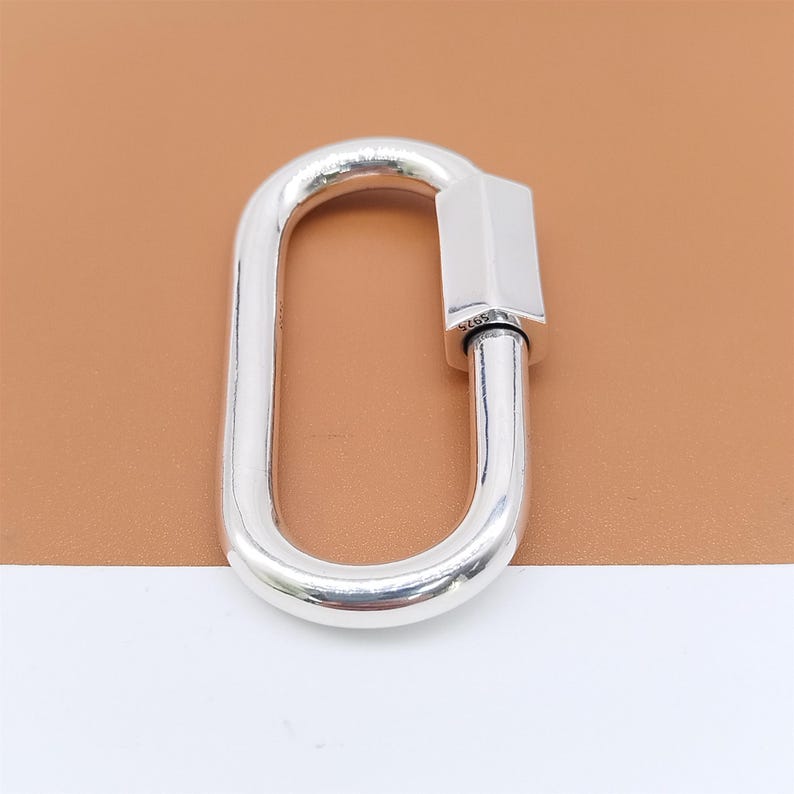 May include: A polished silver oval carabiner clasp. The clasp has a rectangular bar on one end and a hinged, curved body. The background is a light brown color.