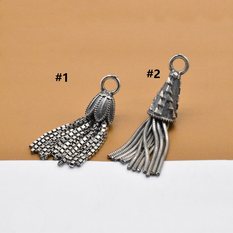 May include: Two silver tassel charms, one with a flower-shaped top and the other with a cone-shaped top. The charms are labeled #1 and #2.