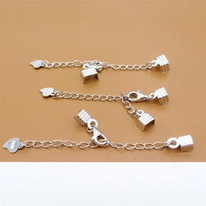 May include: Silver chain extenders for jewellery, featuring lobster clasps and heart-shaped ends. These extenders are designed to lengthen necklaces or bracelets. The background is a plain tan colour.