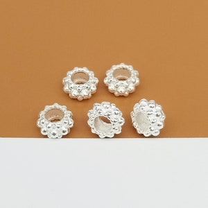 May include: Five silver-colored beads with a floral design. Each bead has a central hole and is surrounded by small, round, textured details. The beads are displayed against a brown and white background.