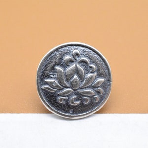 Könnte beinhalten: Ein silberfarbener Metallknopf mit einem erhabenen Design einer Lotusblume.