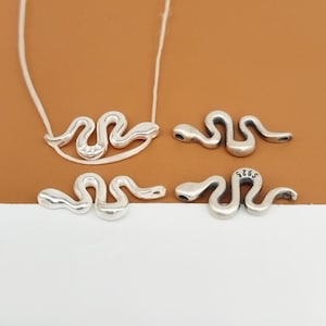 May include: Four silver snake charms, two of which are attached to a white cord necklace. The charms are detailed with a textured snake design.