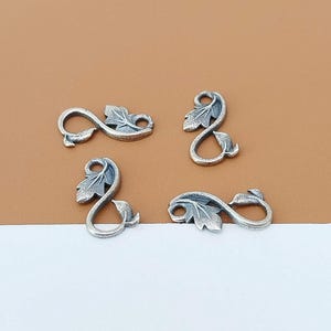 Op de afbeelding: Vier zilverkleurige metalen bedels, elk gevormd als een oneindigheidssymbool met een geïntegreerd bladmotief. De bedels hebben een geoxideerde afwerking die de textuurdetails van de bladeren en de gebogen lijnen van de lussen benadrukt.