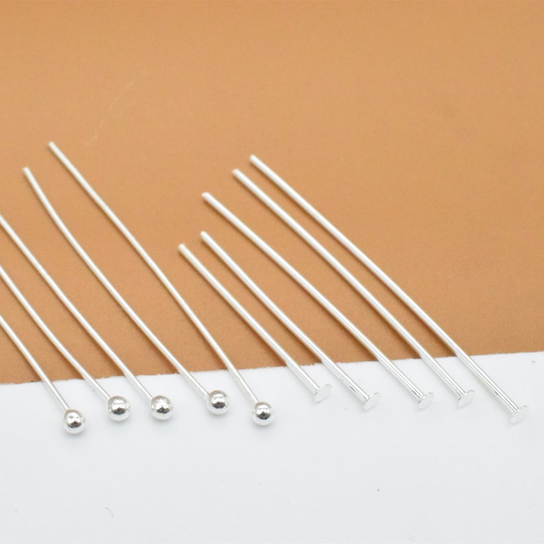 May include: A set of silver head pins with round and flat heads. The pins are arranged in a row on a white surface.