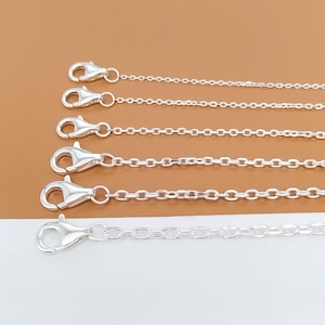 May include: Close-up of five silver-colored chain necklaces with lobster claw clasps. The chains are made of small, interconnected links. The clasps are open, ready to be attached. The background is a light beige color.