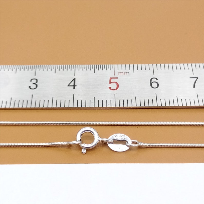 May include: A silver snake chain necklace with a lobster clasp and a small tag that says "925". The necklace is measured against a ruler, showing it is approximately 5 cm long.