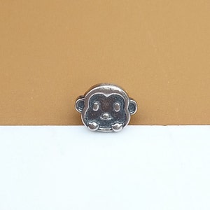 May include: A silver charm shaped like a monkey's face. The charm has a cartoon-like design with large eyes and a small mouth.