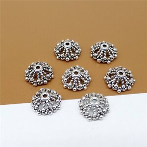 May include: Seven silver-toned metal bead caps with a decorative, intricate design. The caps have a round shape with a hole in the center.