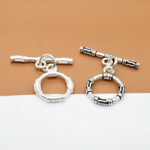 May include: Two silver-colored toggle clasps. Each clasp features a ring with a decorative pattern and a bar. The clasps are designed for jewelry making and are displayed against a brown and white background.