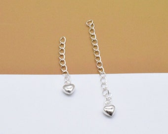 May include: Two silver chains with heart-shaped charms hanging from them. The chains are against a brown and white background.