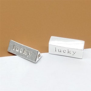 May include: Two silver charm beads with the word "lucky" engraved on them. One bead is rectangular and the other is triangular.