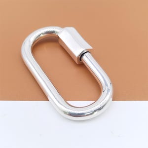 May include: A shiny, silver-colored oval carabiner clasp. The clasp has a rectangular locking mechanism and a smooth, polished finish. The background is a mix of brown and white.