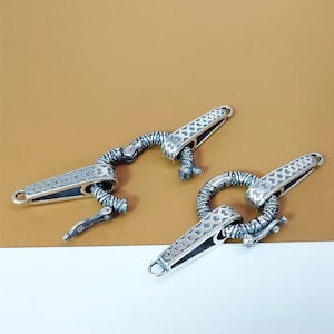 Könnte beinhalten: Zwei silberfarbene Metallverschlüsse mit aufwendigen geometrischen Mustern. Jeder Verschluss hat einen rechteckigen Stab mit einem dekorativen Design und einem gebogenen, beweglichen Arm. Die Verschlüsse sind für die Schmuckherstellung oder ähnliche Handwerke konzipiert.