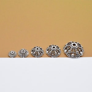 May include: A selection of silver-toned bead caps, each with an intricate, openwork pattern. The caps, varying in size, have a central hole and decorative detailing. They are displayed in a line, highlighting their graduated dimensions.