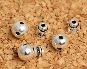May include: Five silver beads, some round and some with a cone shape. The round beads have two holes, and the cone-shaped beads have a single hole. The cone-shaped beads have a black band around the top.