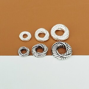May include: Five silver-toned metal rings with a textured design. The rings are different sizes and are arranged in a row on a brown and white surface.