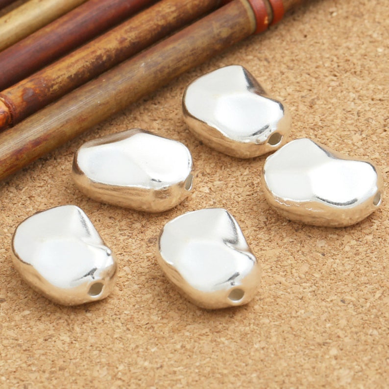 May include: Five silver-colored, irregularly shaped beads with a hole in the center. The beads are arranged in a semi-circle on a brown surface.