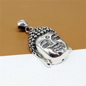 May include: Silver pendant featuring a detailed, three-dimensional depiction of the face of Buddha. The pendant has a loop at the top for attaching to a chain.