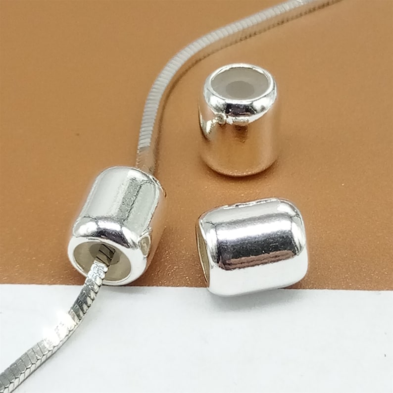 Op de afbeelding: Close-up van drie zilverkleurige cilindrische kralen. E&eacute;n kraal is geregen op een zilveren slangenketting. De kralen hebben een glad, reflecterend oppervlak en een holle binnenkant. De achtergrond is lichtbruin.