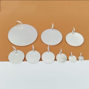 Könnte beinhalten: Eine Sammlung silberfarbener, runder Anhänger in verschiedenen Größen, jeweils mit einer kleinen Schlaufe zum Aufhängen. Die Anhänger sind auf einer hellbraunen Oberfläche angeordnet, was ihre reflektierende Qualität und ihr schlichtes Design hervorhebt.