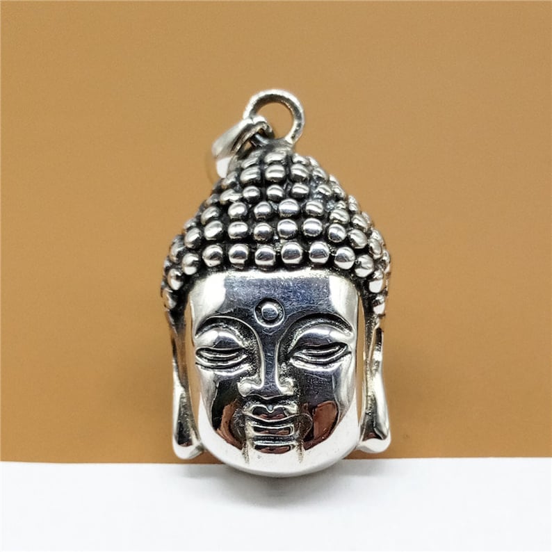 May include: Silver pendant featuring a detailed Buddha head with closed eyes. The pendant has a textured surface and a loop for attaching a chain.
