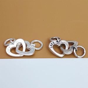 May include: Two silver charm pendants in the shape of interlocking hearts. Each heart has a small loop at the top for attaching to a bracelet or necklace. The charms are marked with the number "925", indicating they are made of sterling silver.