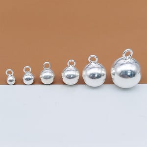May include: Six silver-coloured round beads with loops, varying in size from small to large. The beads are arranged in a row against a brown and white backdrop. These beads are likely for jewellery making and are made of a reflective material.