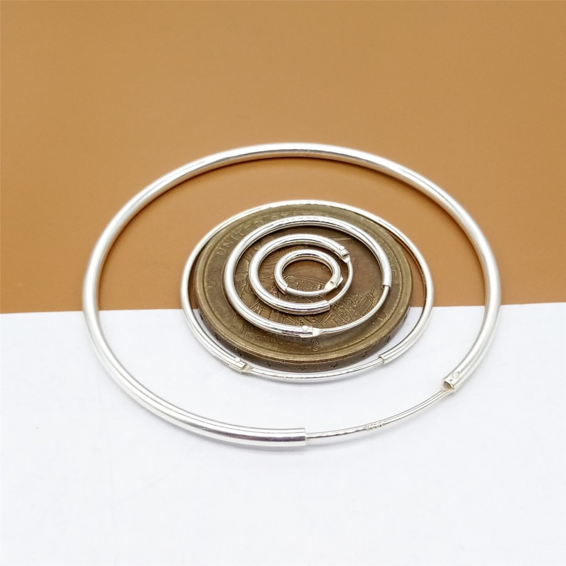 May include: A set of five silver hoop earrings of varying sizes. The earrings are arranged in a circle on a brown and white surface.