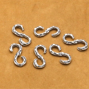 May include: Eight silver-toned metal S-shaped hooks with a textured surface. The hooks are arranged in a random pattern on a brown surface.