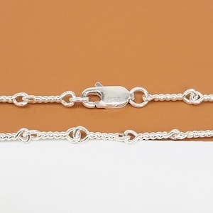 May include: A close-up of a silver necklace with a lobster clasp. The necklace features a delicate chain with knot-like details. The background is a solid, warm brown color.