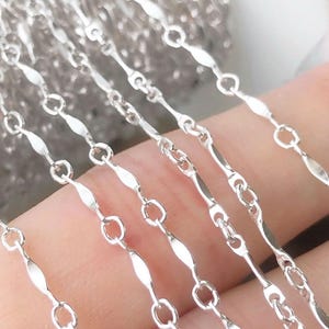 Könnte beinhalten: Nahaufnahme einer silbernen Kette mit einem sich wiederholenden Muster aus ovalen und länglichen Gliedern. Die Kette besteht aus einem glänzenden, reflektierenden Metall, wobei die Glieder durch kleine, kreisförmige Ringe verbunden sind. Wahrscheinlich für die Schmuckherstellung.