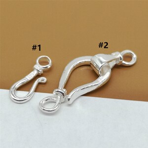 May include: Two silver-colored jewelry clasps are displayed against a neutral background. One is a small hook clasp, and the other is a larger, curved clasp with a ring at each end. The clasps are labeled #1 and #2.