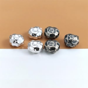 Könnte beinhalten: Sechs silberne und dunkelgraue Metallperlen mit einer strukturierten, unregelmäßigen Oberfläche. Die Perlen sind kugelförmig und haben ein narbiges Aussehen, das an kleine, verwitterte Steine erinnert. Die Perlen werden vor einem braunen und weißen Hintergrund gezeigt.