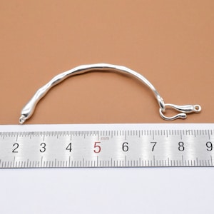 May include: A silver bracelet with a curved, organic shape and a clasp. The bracelet is measured against a ruler, showing it is approximately 5 inches long.