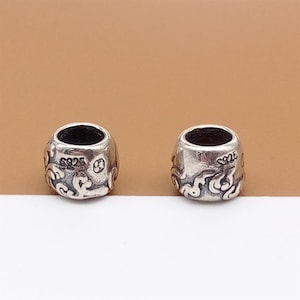 May include: Two silver beads with a cylindrical shape. Each bead has an open centre and is decorated with a swirling pattern and the text "S925". These beads are likely for jewellery making.