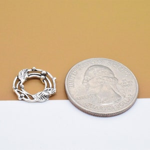 May include: A silver-toned metal wreath charm with a delicate leaf and berry design. The charm is about the size of a US quarter.
