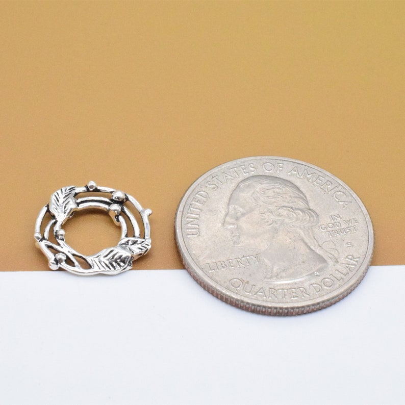 May include: A silver-toned metal wreath charm with a delicate leaf and berry design. The charm is about the size of a US quarter.