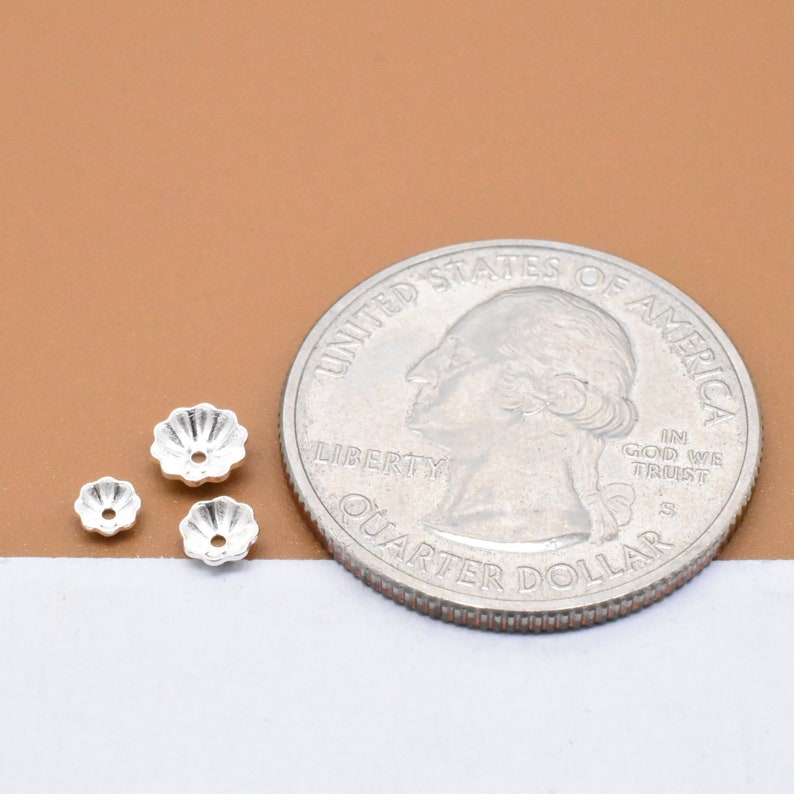 Op de afbeelding: Drie zilverkleurige metalen bloemvormige kralencaps, elk met een klein gaatje in het midden, worden getoond naast een Amerikaanse kwart dollar munt voor groottevergelijking.