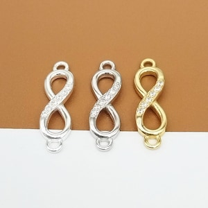 Pode incluir: Três pingentes de charme de infinito em tons de prata e ouro. Cada pingente tem a forma do símbolo do infinito, com uma fileira de pequenos cristais transparentes no centro. Os pingentes têm pequenas argolas na parte superior e inferior.
