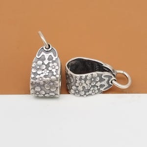 May include: Two silver-toned metal pendant bails with a floral design. The bails are oval shaped and have a small loop at the top for attaching a pendant.