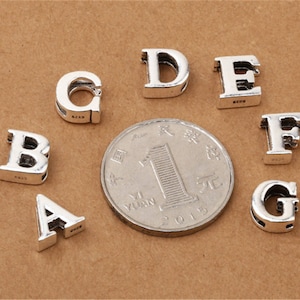 May include: Silver charm beads in the shape of the letters A, B, C, D, E, F, and G. The beads are arranged on a brown surface.