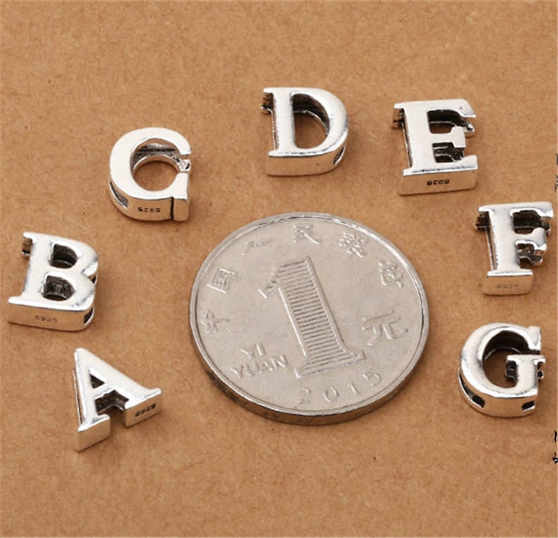 May include: Silver charm beads in the shape of the letters A, B, C, D, E, F, and G. The beads are arranged on a brown surface.