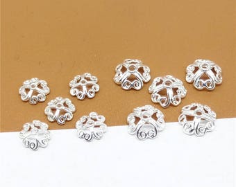 May include: Ten silver-toned metal bead caps with a decorative, floral design. The caps are arranged on a brown surface.
