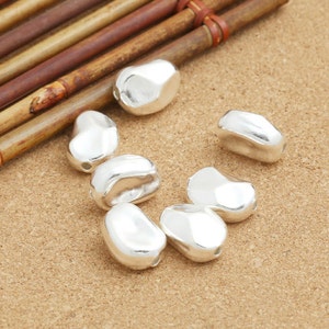 May include: Seven silver-colored, bean-shaped beads, with a smooth, slightly textured surface. The beads are arranged on a light brown, textured surface.