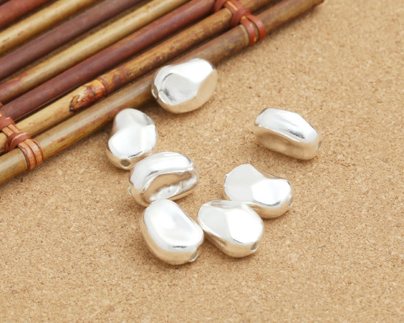 May include: Seven silver-colored, bean-shaped beads, with a smooth, slightly textured surface. The beads are arranged on a light brown, textured surface.